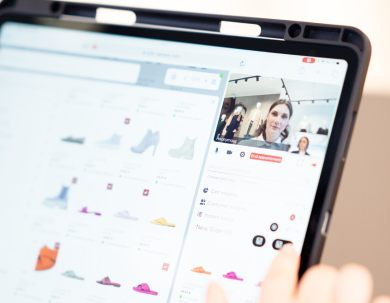 Eine Person stöbert nach Schuhe in einem Onlineshop auf einem Tablet, mit Produktbildern auf der linken Seite und einem Videoanruf auf der rechten Seite. Der Finger der Person zeigt auf eine Option auf dem Bildschirm.
